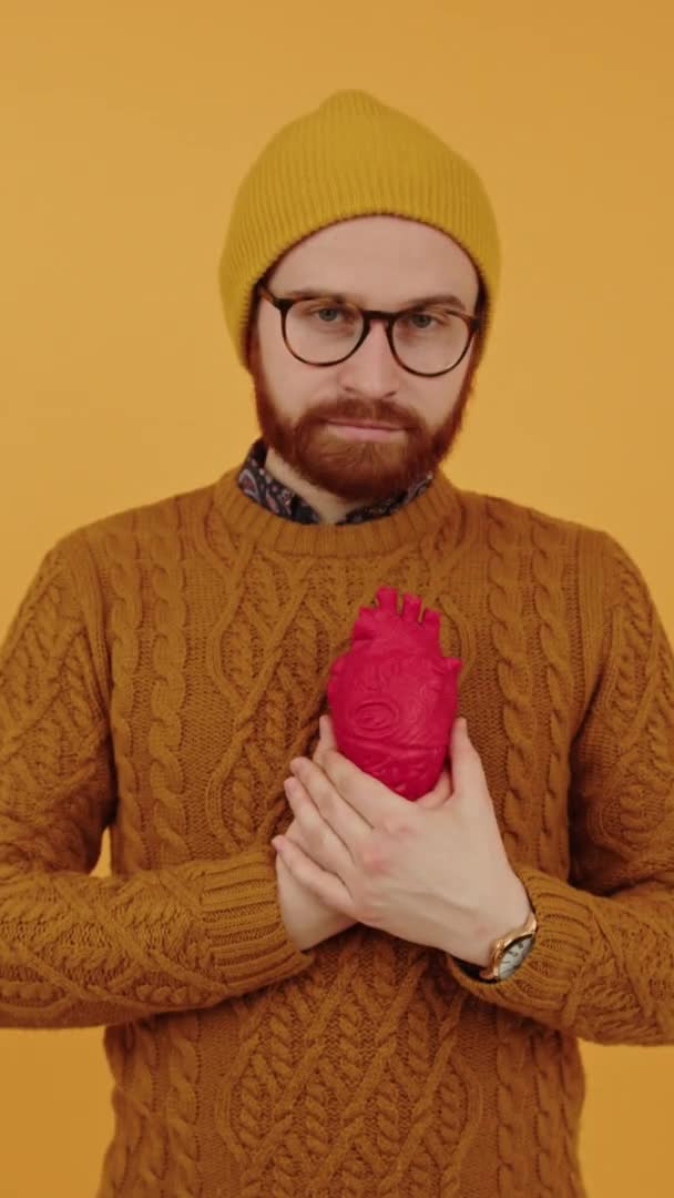 Vertical shot of a serious caucasian bearded millennial guy in eyewear and a beanie holding red artificial heart in front of his chest over yellow background. Health concept. — Stock Video