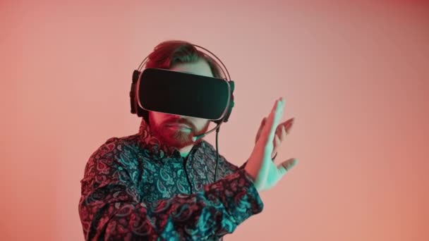 Concepto VR. Milenial caucásico barbudo hombre en una camisa estampada con gafas VR profesionales y auriculares sobre fondo rosa. — Vídeo de stock