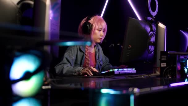 Konsantre olmuş pembe saçlı Asyalı kız büyük kulaklıklar takıyor ve kot ceket giyiyor. Oyun yarışmasının ortasında bilgisayar ekranına bakıyor.. — Stok video