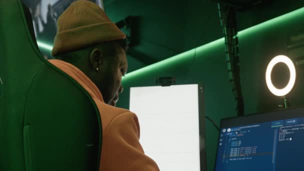 Вид сзади на африканского американца в оранжевой толстовке и шляпе, сидящего на высоком стуле, смотрящего на экран компьютера. — стоковое видео