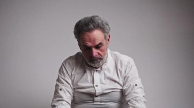 Stüdyoda, 60 'lı yaşlarda, üzgün, beyaz saçlı, başını eğmiş, ara sıra kameraya bakan bir adamın görüntüleri var. Gri arkaplan.