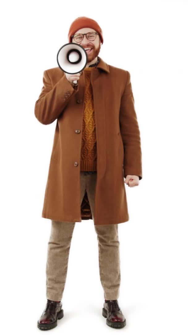 Vertical full body shot of a caucasian bearded elegant hipster man in brown autumn clothes cheerfully shouting via megaphone over white background. — Stock Video