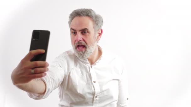 Смішний старий сіро-волохатий підприємець бере селфі зі своїм чорним смартфоном і робить дурні обличчя на білому тлі . — стокове відео