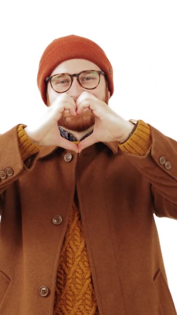 Вертикальная студия снимает молодого бородатого мужчину в осенней одежде и очках, показывающих жест сердца перед камерой. Романтическая концепция. — стоковое видео