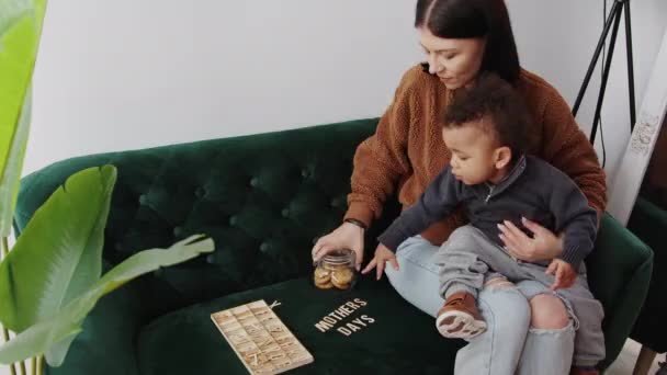 Mor öppnar en burk med kakor till sin son medan du spelar ett vokabulär spel i vardagsrummet kopiera utrymme medium full shot — Stockvideo