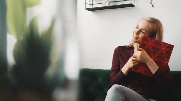 Uma jovem mulher branca loira excitada sentada em um sofá em um apartamento, segurando um presente embrulhado e abraçando-o. — Vídeo de Stock