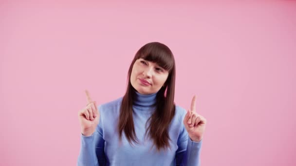 Positiv kaukasisk brunett med lugg i babyblå polotröja som pekar uppåt med två pekfingrar in i kopieringsutrymmet. Studio skjuten med rosa bakgrund. Reklamförslag. — Stockvideo