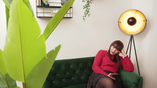 Plan intérieur d'une jeune femme caucasienne en pull rouge assise sur un canapé derrière une plante et utilisant son nouveau téléphone. — Video
