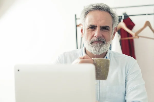 Freelance ondernemer concept. Kaukasisch grijs harige man met baard zittend achter zijn laptop, een kop koffie of thee vasthoudend en kijkend naar camera, geïsoleerd — Stockfoto
