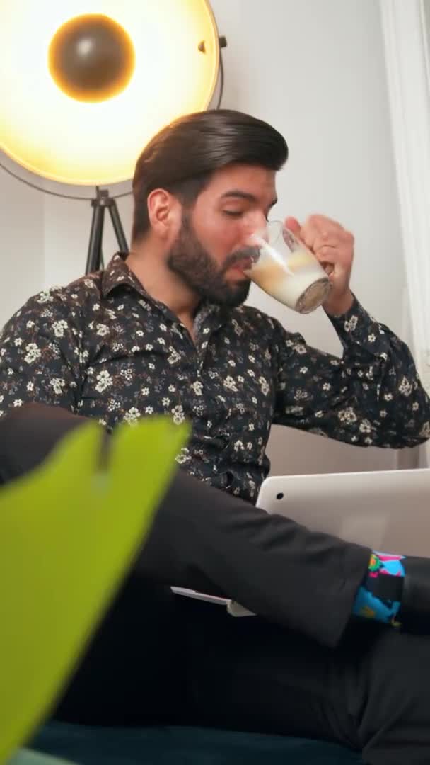 Бородатий красивий чоловік у свої 30 років п'є каву і намагається не сміятися під час відеозапису в своєму домашньому офісі, вертикально — стокове відео