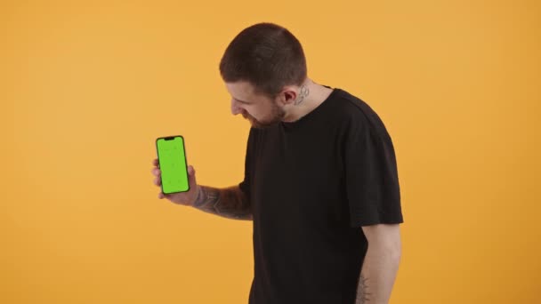 Привлекательный бородатый мужчина держит смартфон с зеленым дисплеем, показывает большие пальцы вверх, глядя на камеру студии снимок — стоковое видео