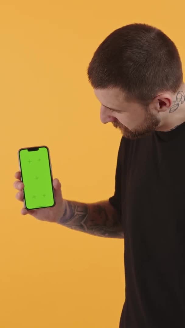 Jovem caucasiano mostrando smartphone com tela verde, olhando para a câmera e mostrando polegares para cima estúdio de vídeo vertical tiro — Vídeo de Stock