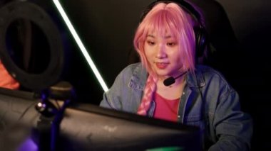 20 'li yaşlarında, pembe saçlı bir Asyalı kadın çevrimiçi bir oyuna katılıyor, bir ekranın önünde oturuyor, kazandıktan sonra tezahürat yapıyor. Yüksek kalite 4k görüntü