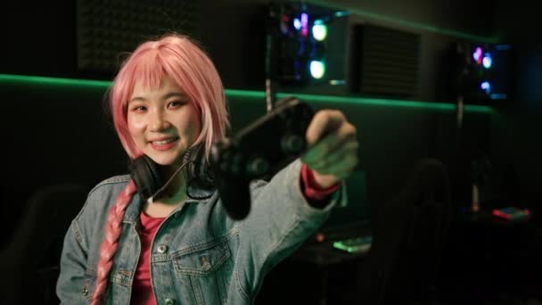 ピンクの長い編組の可愛いアジア系の女の子の暗い部屋で撮影された室内では ゲームパッドを片手に親指を立ててデニムジャケットを身に着けていました 高品質4K映像 — ストック動画