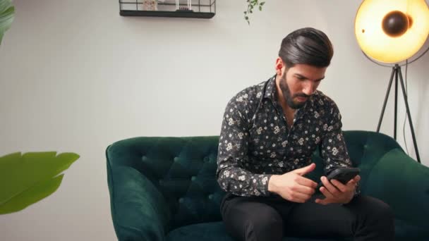 Латиноамериканец Бородой Сидит Зеленом Диване Смотрит Свой Мобильный Телефон Аплодирует — стоковое видео