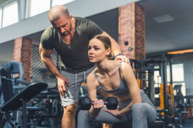 Sportif Avrupalı kadın spor salonunda halterle omuz egzersizi yaparken antrenörü duruşunu düzeltiyor..