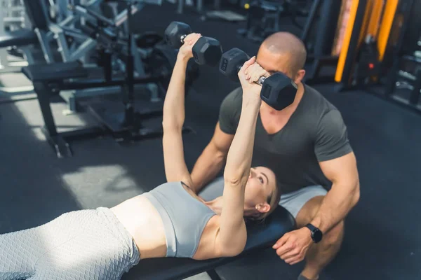 Gym activities. Wellbeing and physical health concept. Two black dumbbells lifted by a young European girl in a gray gym clothes set.