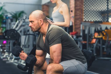 Kılsız, sakalsız, güçlü bir erkek vücut geliştiricinin halter boyunca ağırlık kaldırırken, iyi donanımlı bir spor salonunda bir bankta otururken profil çekimi. Yüksek kalite fotoğraf