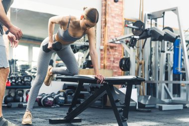 Çekici, ince yapılı, esmer bir kadın spor salonundaki bir bankta eğiliyor, koluyla dambılı kaldırıyor. Solda duran tanınmayan beyaz bir adam var. Yüksek kalite fotoğraf
