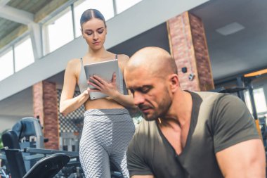 Orta yaşlı kel seksi erkek vücut geliştirici oturup ağırlık kaldırıyor. Genç, beyaz, çekici bir kadın üzerinde duruyor ve tabletteki sonuçları kontrol ediyor. Yüksek kalite fotoğraf