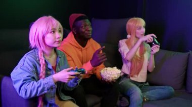 Oyun odasındaki kanepede oturan üç farklı ırktan arkadaş. Video oyunu oynayan iki kız, galibiyetten sonra tezahürat yapan, turnuvada oynayan. Yüksek kalite 4k görüntü
