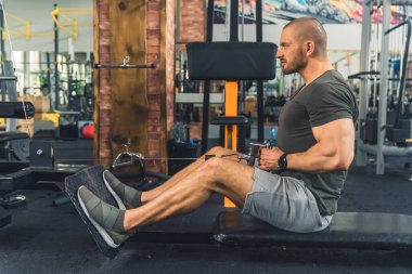 Kel, uzun boylu, Avrupalı bin yıllık bir adam kürek çekme makinesi egzersizi sırasında bir bankta oturup kaslarını sıkıyor. Yüksek kalite fotoğraf