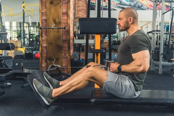 Bald tall european millenial man sitting on a bench tightening his muscles during an exercise on a rowing machine. High quality photo