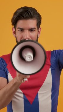 Beyaz sakallı, çekici, aktif ve mutlu 20 'li yaşlarda Küba bayrağı tişörtü giyen, sarı arka plan stüdyo portresi üzerinde dikey olarak izole edilmiş, hoparlörden konuşan genç bir adam. Yüksek kalite 4k görüntü
