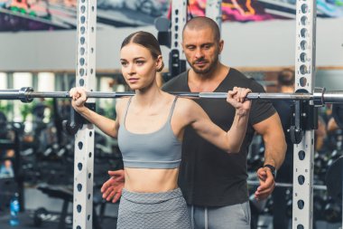 Omuz halteri kaldırma. Erkek kişisel antrenörünün rehberliğinde halter deneyen kendine güvenen beyaz bir kız. Spor salonunun içi. Yüksek kalite fotoğraf