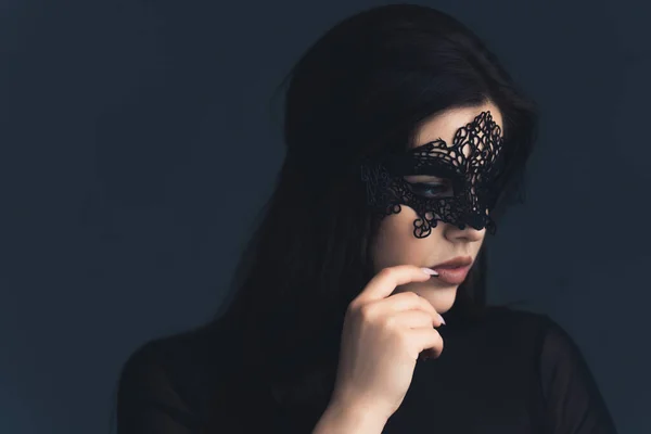 Close-up studio shot over dark blue background of a dark-haired seductive beautiful caucasian woman wearing black clothes and a black lacy mask, touching her face with her finger. High quality photo