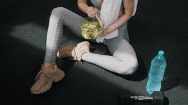 Gri taytlı, elinde bir kase yeşil salata tutan, spor salonunun zemininde oturan atletik, rahat, beyaz bir kadın. Sağlıklı yaşam tarzı kavramı. Yüksek kalite 4k görüntü