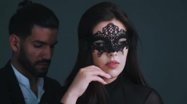 Siyah elbiseli, siyah dantelli karnaval maskeli seksi beyaz kadın kameraya bakıyor, yüzüne dokunuyor ve yakışıklı sevgilisinin önünde duruyor. Karanlık arka plan. Stüdyo çekimi. Yüksek