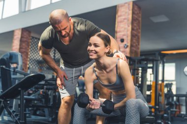 Spor salonu konsepti. Bir bankta otururken dambılı kaldıran beyaz kadın gülümsüyor çünkü artık güçlü ve güçlü bir kadın olduğunu görüyor. Erkek özel antrenör arkada. Yüksek kalite fotoğraf