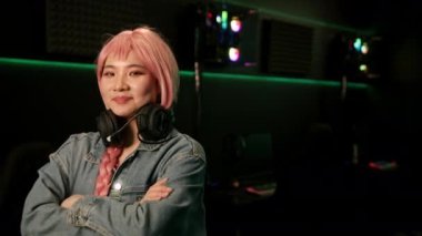 Pembe peruklu, elinde siyah oyun kumandası olan ve gülümseyen kulaklıkla fotoğrafa poz veren sevimli Asyalı kız. Yüksek kalite 4k görüntü