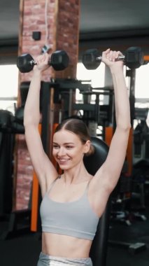 Otururken dambılları kaldıran güçlü, beyaz, genç bir kadın vücut geliştirme ustasının kapalı alanda tam vücut çekimi. - Evet. Yüksek kalite 4k görüntü