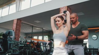 20 'li yaşlarda güzel, koyu saçlı, zayıf bir kadın bir bankta oturuyor ve sırtı düz ağır bir dambılı kaldırıyor. Erkek kişisel koçu arkasında duruyor ve yardım ediyor. Yüksek kalite 4k görüntü