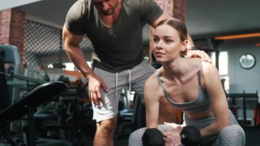 Sportif Avrupalı kadın spor salonundaki bir bankta halterle omuz egzersizi yaparken kişisel koçu duruşunu düzeltiyor. Yüksek kalite 4k görüntü