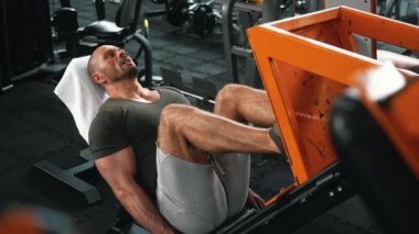 Son derece yorgun, kel, yapılı bir adam bankta uzanmış, ayak presi üzerinde egzersiz yapıyor. İyi donanımlı bir spor salonunda kapalı bir atış. Yüksek kalite 4k görüntü