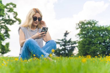 Sarışın, beyaz bir kız çimlerin üzerinde oturuyor ve cep telefonunu açık havada kullanıyor. Yüksek kalite fotoğraf