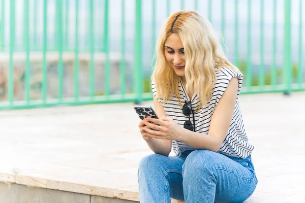Sarışın, beyaz bir kız akıllı telefondan mesaj okuyor ve gülümsüyor. Açık havada orta boy. Yüksek kalite fotoğraf