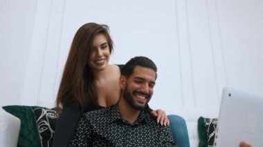 30 'lu yaşlarındaki neşeli sakallı latin adam elinde dizüstü bilgisayar, arkasında kanepede oturan esmer, mutlu kadın. Yüksek kalite 4k görüntü