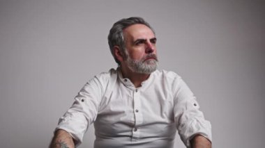 50 'li yaşlarda, beyaz saçlı, ciddi bir ifadeyle yüzünü yana çevirmiş, sonra kapalı alanda çekilmiş, gülümseyen gri arka plan stüdyosuna bakıyor. Yüksek kalite 4k görüntü