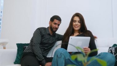 Uzun saçlı güzel kız arkadaşıyla kanepede oturan desenli siyah gömlekli yakışıklı Kübalı adam. Modern apartman dairesi. Yüksek kalite 4k görüntü