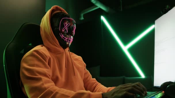 匿名互联网身份的概念 穿着黄色帽衫的人在一台程式设计的计算机前 戴着恐怖的发光霓虹灯面罩 看着相机 高质量的4K镜头 — 图库视频影像