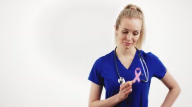Boynunda steteskop olan gururlu ve kendine güvenen beyaz kadın doktor ya da hemşire kalbine pembe bir kurdele takarak beyaz arka plandan kameraya bakıyor. Yüksek kalite 4k görüntü