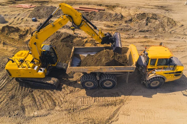 A yellow huge digger loading sand onto a long tipper truck. Works at a road construction site. Drone photo. High quality photo