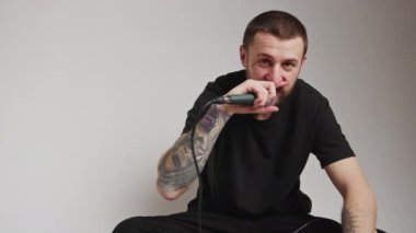 Sakallı, beyaz adam rap yapıyor ve mikrofonu kameraya doğrultuyor. Gri arka plan, stüdyo görüntülerini izole ediyor. Yüksek kalite 4k görüntü