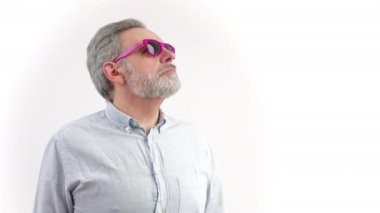 50 'li yaşlarda, sakallı, gri saçlı bir adam gözlerini kaçırıyor ve pembe güneş gözlüğünü çıkarıyor. Beyaz arka plan stüdyosu içeride fotokopi çekiyor. Yüksek kalite 4k görüntü