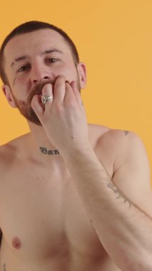 Dövmeli üstsüz adam bıyığına dokunuyor ve dikey video sarı arka plan çekimine gülümsüyor. Yüksek kalite 4k görüntü