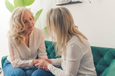 40 'lı yaşlarda, koyu yeşil bir kanepede oturmuş el ele tutuşan iki çekici sarışın kadın. Yüksek kalite fotoğraf
